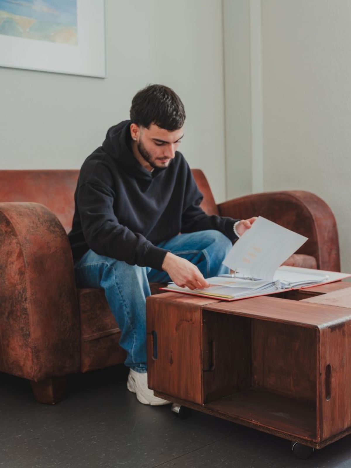 Junger Mann sitzt auf einem Sessel und blättert konzentriert durch einen Ordner auf einem Tisch vor ihm.