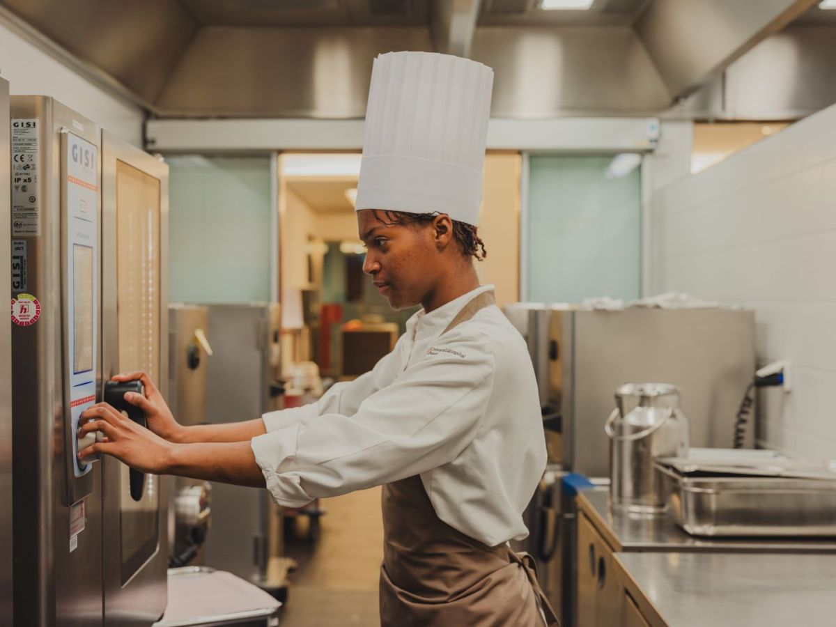 Köchin in Arbeitskleidung und mit Kochmütze bedient konzentriert ein Küchengerät in einer professionellen Grossküche.
