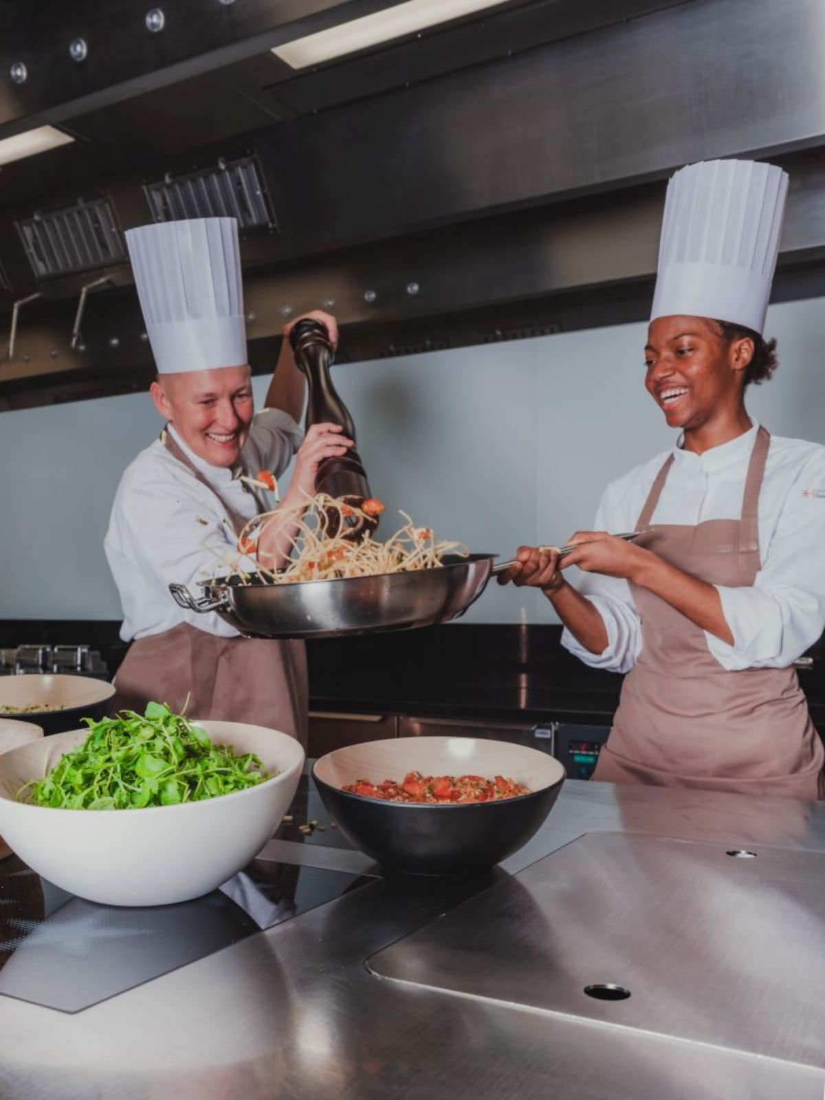 „Zwei Köchinnen stehen in einer Grossküche und bereiten gemeinsam ein Gericht zu; eine schwenkt eine Pfanne mit Pasta, während die andere sie hält, beide lachen.