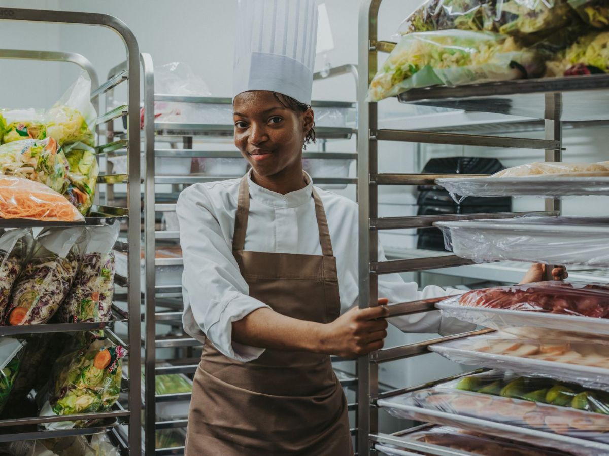 Köchin in Arbeitskleidung steht zwischen Regalen mit vorbereiteten Lebensmitteln und schiebt ein Gestell mit Zutaten in einer Grossküche.