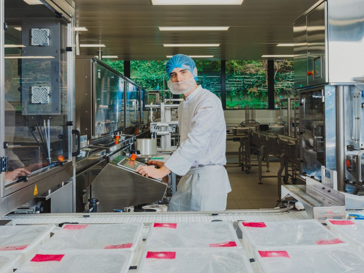 Mitarbeiter in Schutzkleidung mit Haarnetz und Mundschutz steht in einer Produktionshalle und bedient eine Anlage; im Vordergrund stehen verpackte Produkte auf einem Förderband.