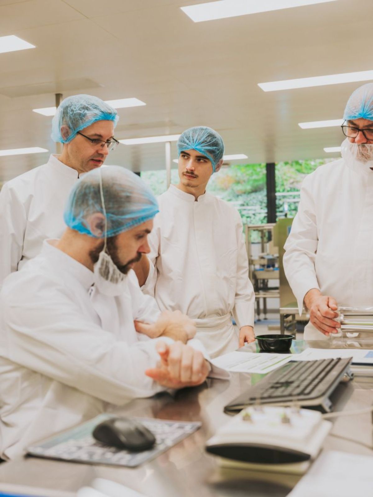 Vier Mitarbeitende in Schutzkleidung und mit Haarnetzen besprechen sich an einem Arbeitstisch in einer Produktionsumgebung und schauen gemeinsam auf Unterlagen.