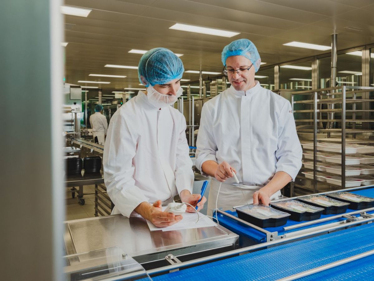 Zwei Mitarbeitende in Schutzkleidung mit Haarnetzen arbeiten an einer Produktionslinie; sie prüfen verpackte Produkte und halten Ergebnisse auf einem Formular fest.