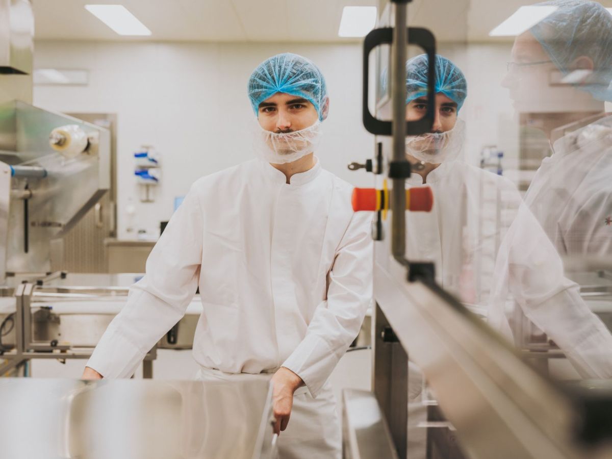 Mitarbeiter in Schutzkleidung mit Haarnetz und Mundschutz steht in einer Produktionsumgebung neben einer Anlage und blickt in die Kamera; seine Spiegelung ist im Glas sichtbar.