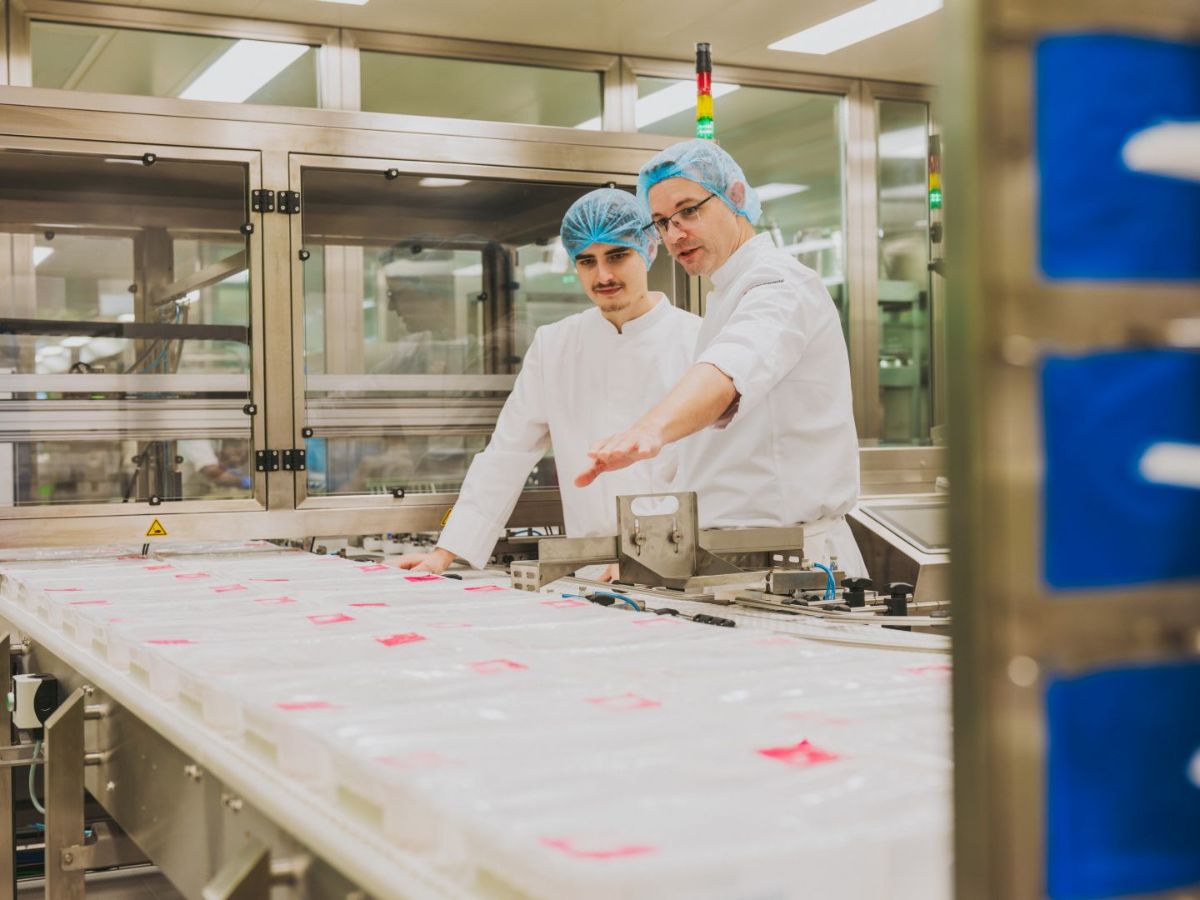 Zwei Mitarbeitende in Schutzkleidung mit Haarnetzen stehen an einer Produktionslinie und überprüfen gemeinsam verpackte Produkte auf einem Förderband.