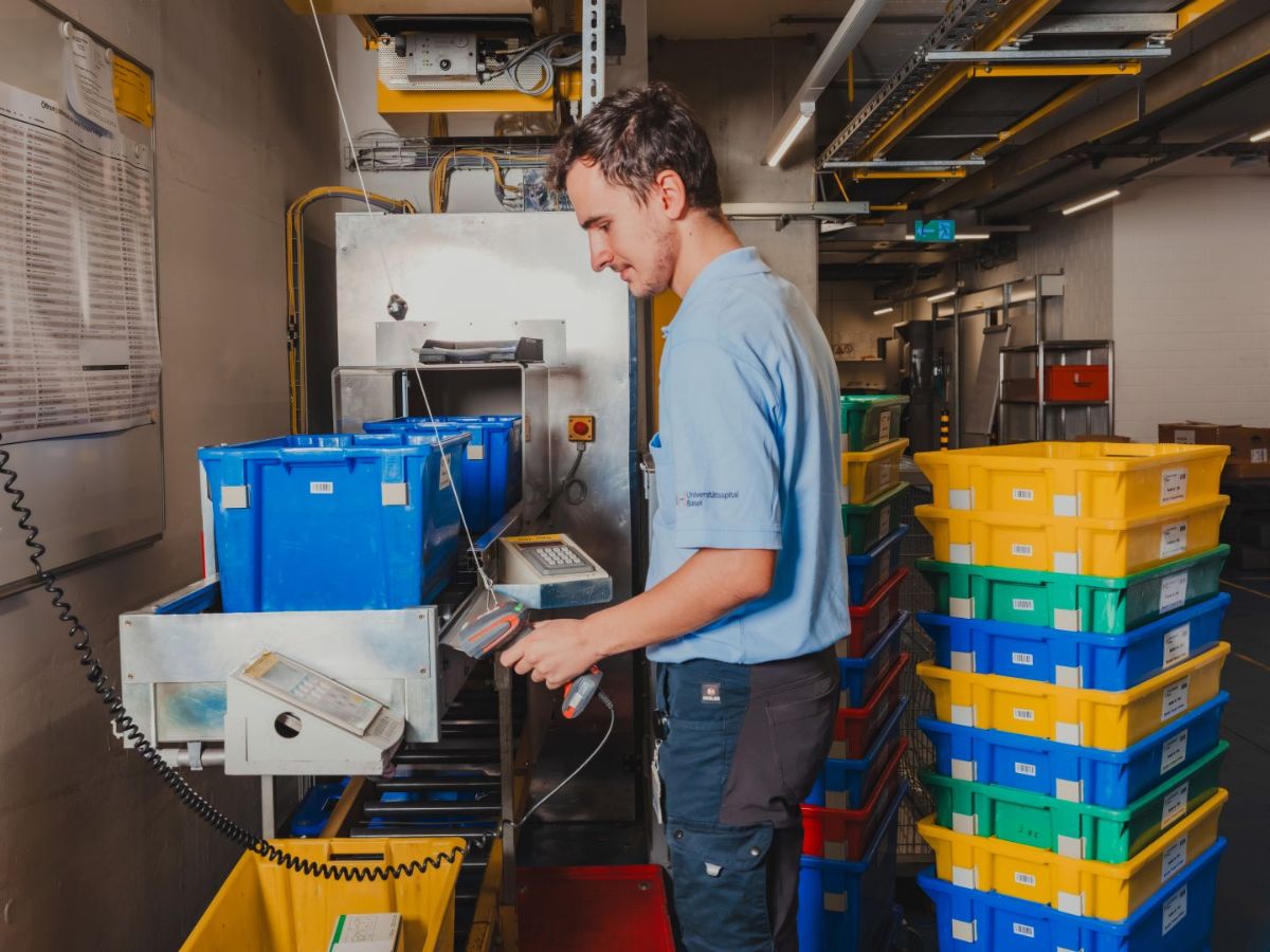 Mitarbeiter in einem Lager scannt Waren mit einem Handscanner; neben ihm stehen gestapelte Transportboxen in verschiedenen Farben.