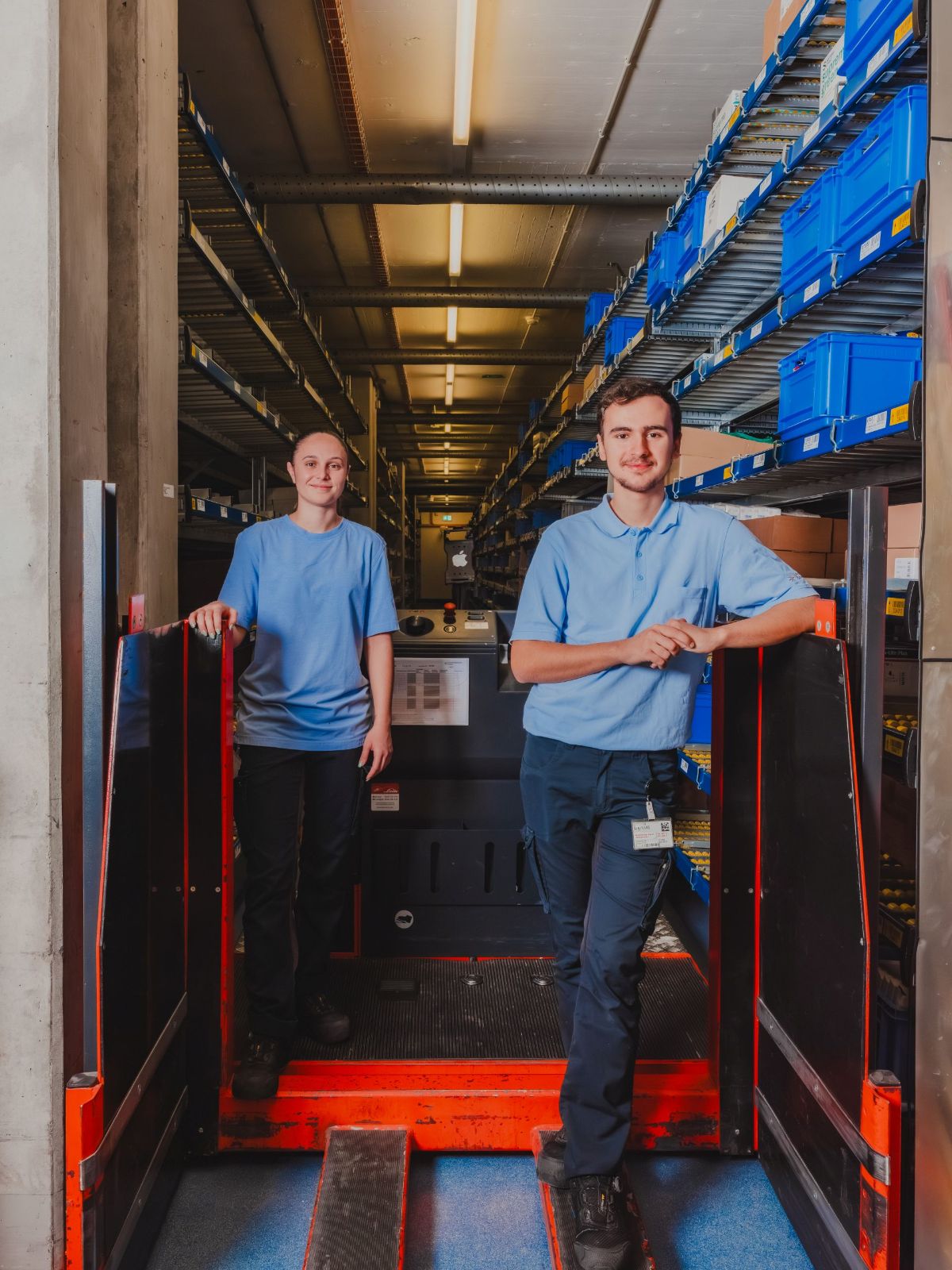 Zwei junge Personen stehen in einem Lager vor einem Kommissioniergerät; im Hintergrund sind Regale mit blauen Boxen zu sehen.