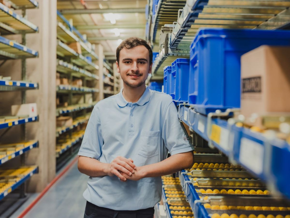Mitarbeiter steht in einem Lagergang zwischen Regalen mit blauen Behältern und blickt in die Kamera.