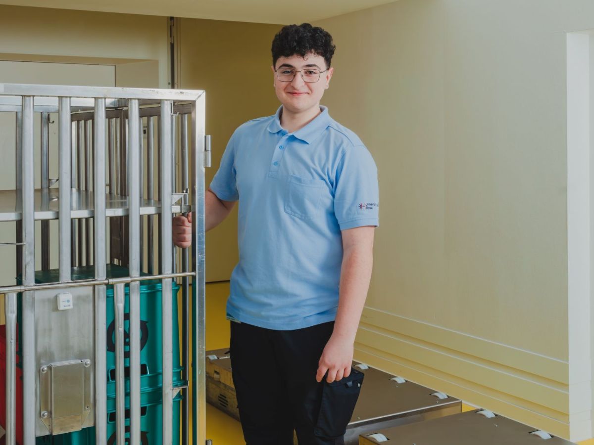 Mitarbeiter steht neben einem Transportwagen mit Behältern in einem Innenbereich und blickt in die Kamera.