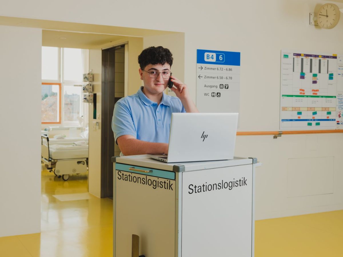 Mitarbeiter der Stationslogistik arbeitet an einem Laptop und telefoniert auf einem Flur im Spital