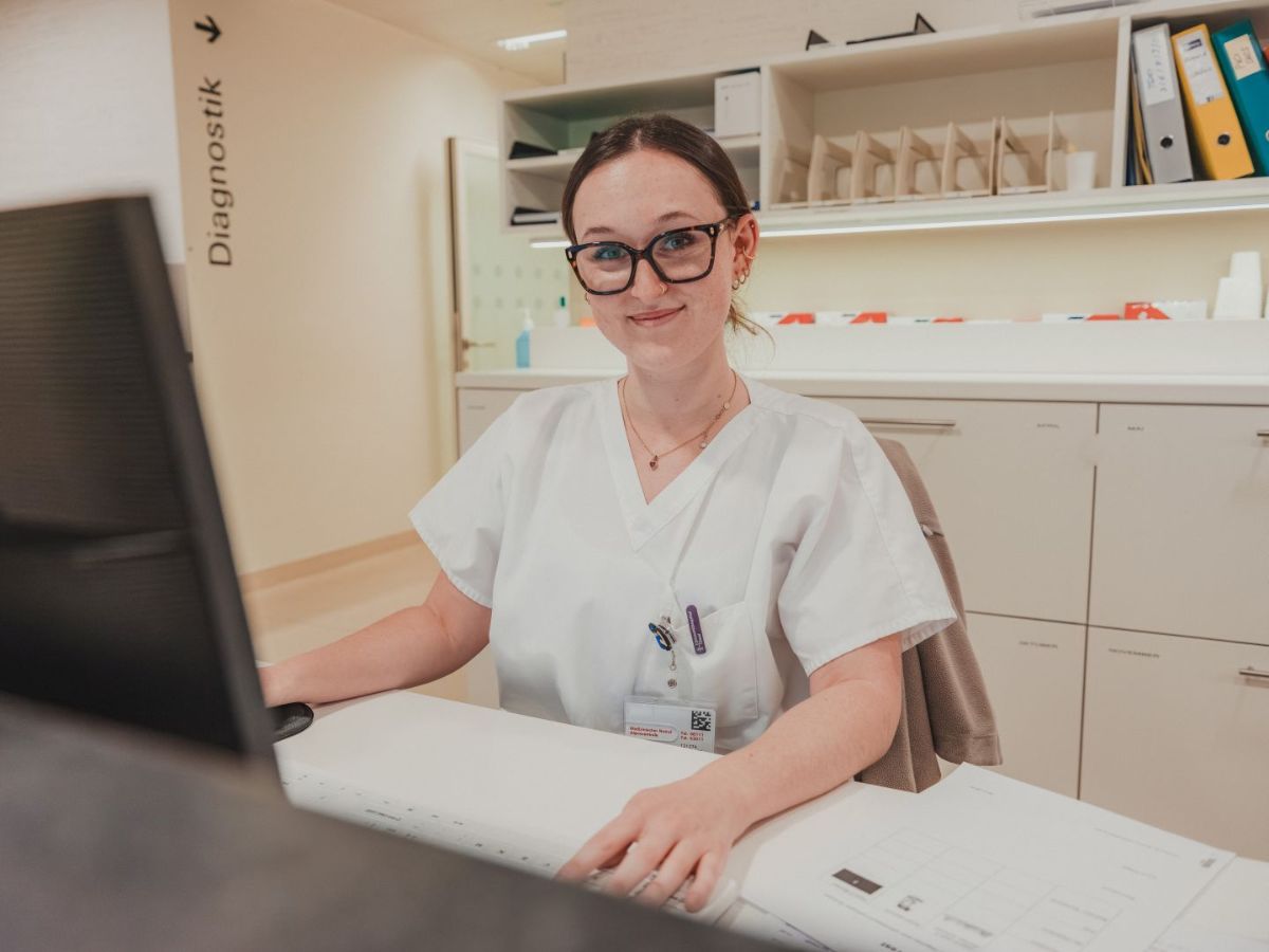 Junge medizinische Mitarbeiterin in heller Arbeitskleidung sitzt an einem Empfangsschalter, lächelt in die Kamera und arbeitet am Computer; im Hintergrund sind Ordnerregale und ein Bereich mit der Beschriftung "Diagnostik" zu sehen.