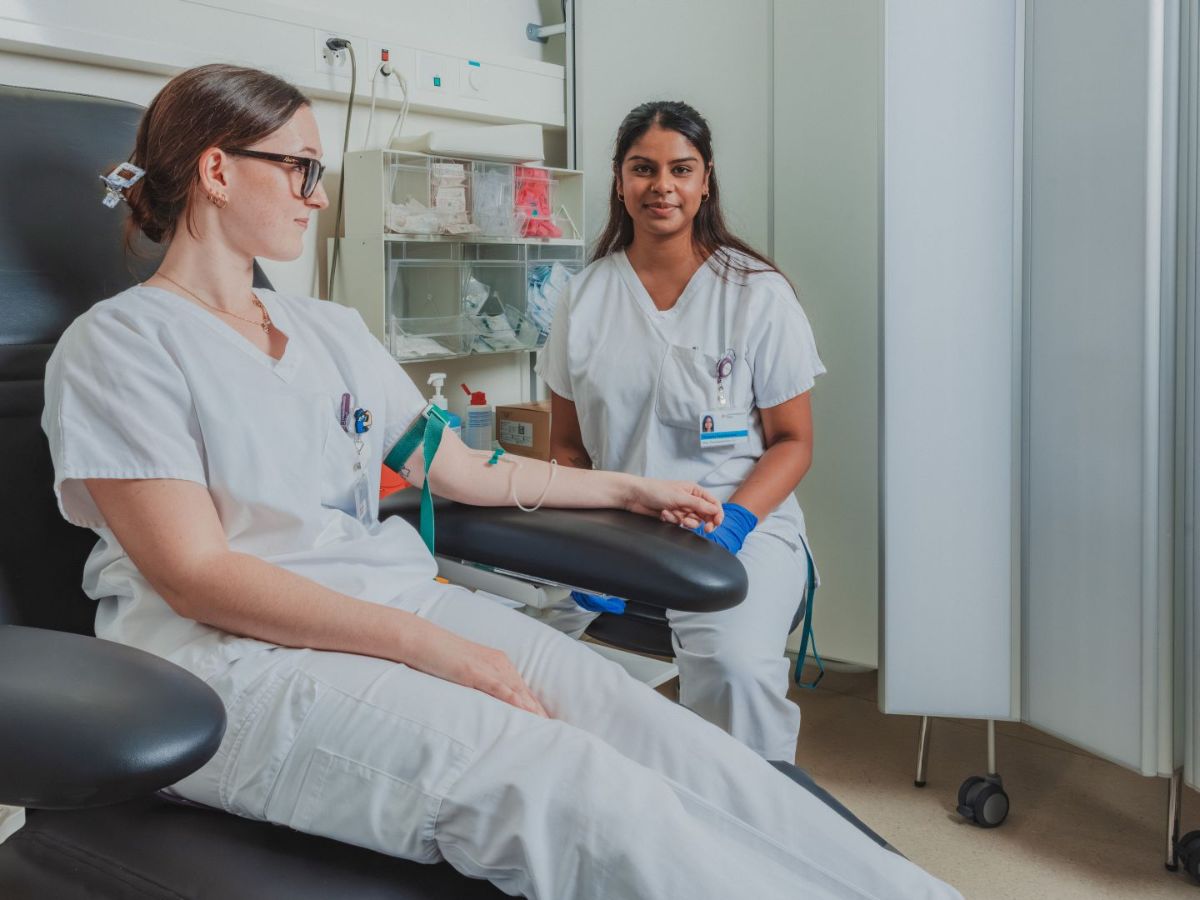 Zwei medizinische Mitarbeiterinnen in heller Arbeitskleidung bei einer Blutabnahme: Eine sitzt entspannt im Behandlungsstuhl mit angelegtem Stauschlauch und Zugang am Arm, während die andere daneben sitzt, Handschuhe trägt und sie betreut; im Hintergrund sind medizinische Utensilien und ein Behandlungsraum zu sehen.