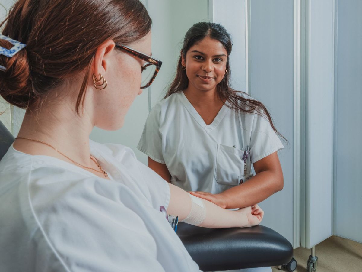 Zwei medizinische Mitarbeiterinnen in einem Behandlungsraum: Eine sitzt im Stuhl mit verbundenem Arm nach einer Blutabnahme, während die andere ihr gegenüber sitzt, lächelt und sich um sie kümmert.