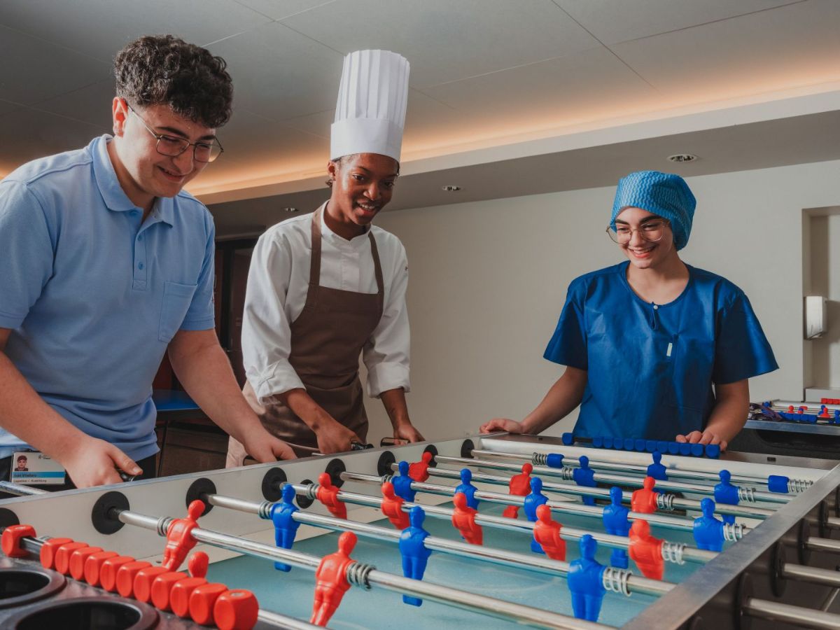 Drei junge Menschen in unterschiedlichen Berufsbekleidungen spielen gemeinsam Tischfussball und lachen.