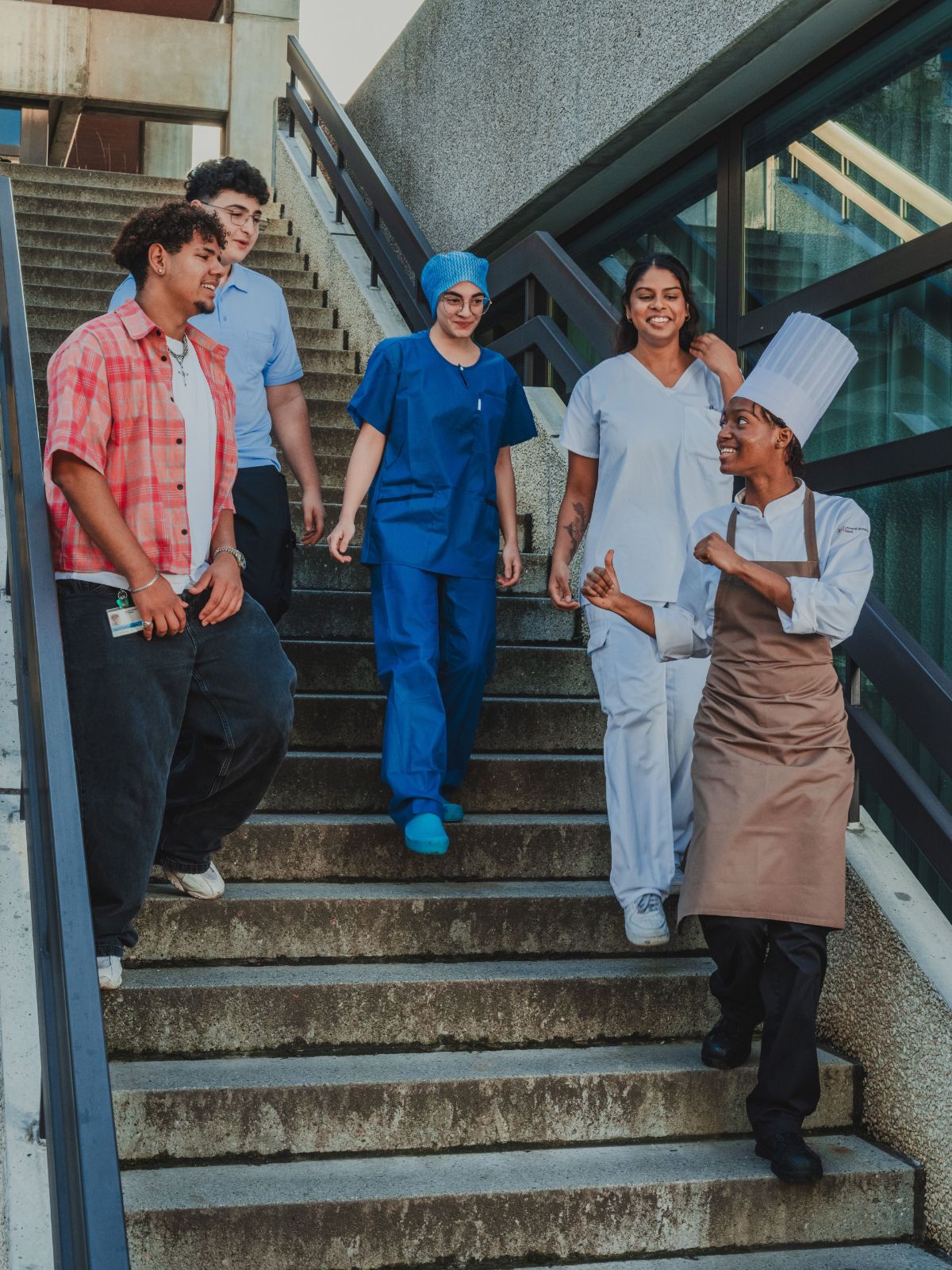 Fünf junge Personen in unterschiedlichen Berufskleidungen gehen gemeinsam eine Treppe hinunter und unterhalten sich; darunter sind Pflegefachpersonen und ein Koch, die in einer lockeren, kollegialen Stimmung unterwegs sind.