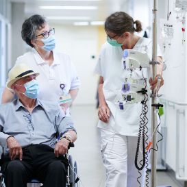 Patient wird von 2 Pflegerinnen im Rollstuhl über Gang gefahren